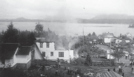 A group of people stand watching smoke billowing from a building. Around the building are more buildings, a street, and trees. 