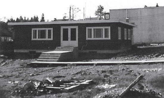 A dark-coloured, one-storey building surrounded by dirt. 