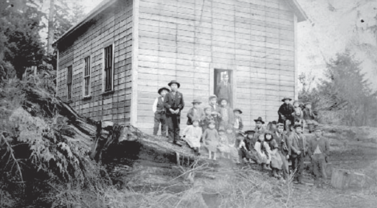 About 20 people, mostly children, stand and sit on a fallen trunk of a tree in front of a building.