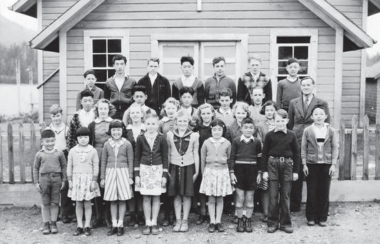 About 30 people, mostly children, pose for the camera, standing in rows with their backs straight. They are in front of a building.