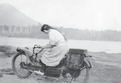 A woman with her hair in a long braid wearing a dress riding a motorcycle along a body of water.