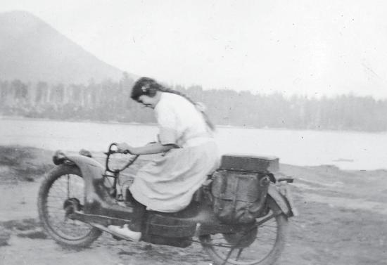 A woman with her hair in a long braid wearing a dress riding a motorcycle along a body of water.