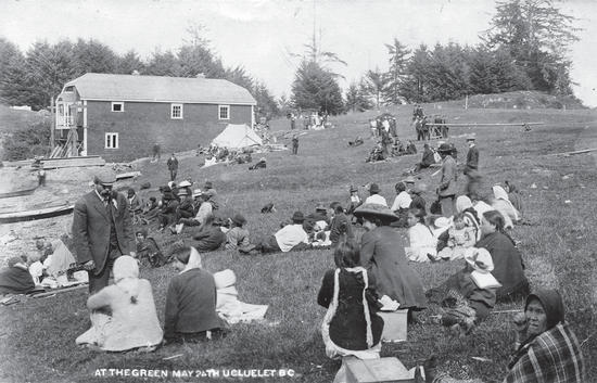 Numerous people sitting and standing on a grassy incline. Behind them is a building and trees. 