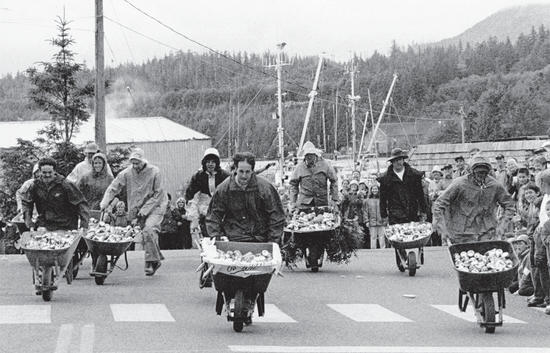 About nine people push filled wheelbarrows on a road. A line of people along the road watches.