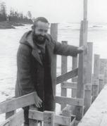A person smiles at the camera while leaning on wooden planks nailed together. Behind them is a beach. 