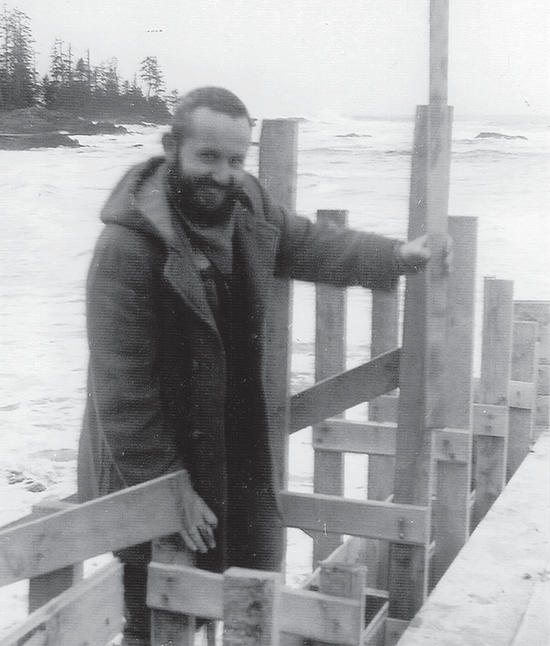 A person smiles at the camera while leaning on wooden planks nailed together. Behind them is a beach. 
