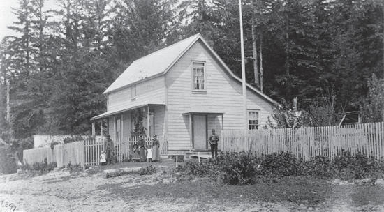 A light-coloured house made of wood in front of a forest. The house is surrounded by fencing, and within the fencing stand seven people. The group is a mixture of adults and children.