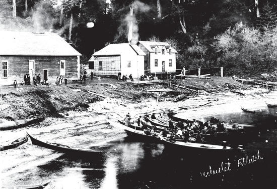 A group of people are sitting on canoes docked at a beach. Behind the beach are two wooden buildings in front of which is another group of people standing and watching the other group. On the photograph, the following words are written: Ucluelet Potlatch