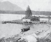 A sailboat docked at a beach. By the beach is a wooden dock with two buildings. In the background are a forest and a mountain.