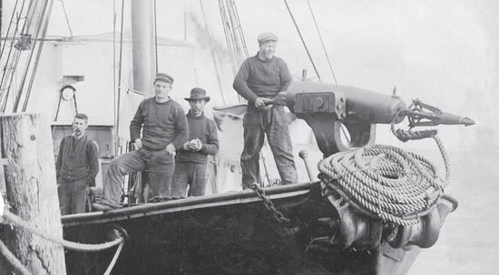 Four men stand at the end of a docked, dark-coloured boat. Three men look at the camera while one man looks away as he handles a harpoon gun. 