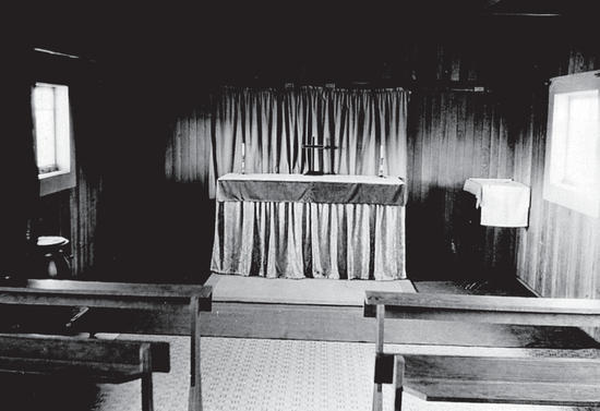 The inside of a small, empty church. Wooden pews are lined up in front of a table covered by a tablecloth and topped with a cross. Behind the stable is a curtain.