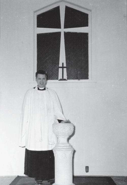 A person in robes smiling at the camera while their left hand rests on a light-coloured structure that reaches their hip. Behind them is a window with a cross in it, and a small cross sitting on its windowsill.