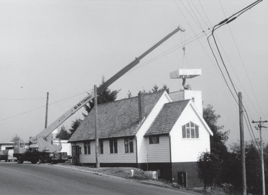 A construction vehicle, a large object hanging from its long arm. It is being lowered behind a light-coloured wooden building.