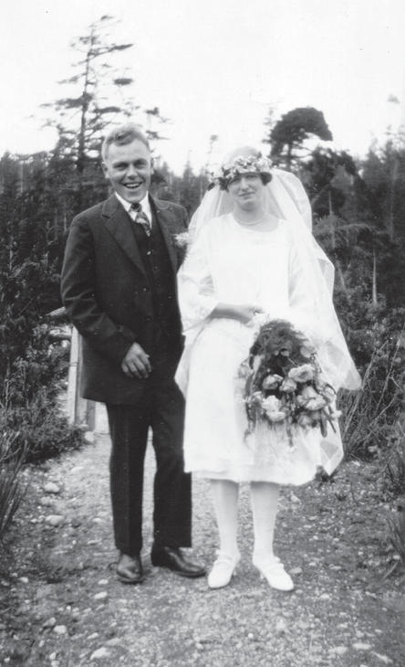 A man in a three-piece suit and a woman wearing a wedding dress and a veil. The woman holds flowers in one hand. They smile at the camera while standing on a dirt pathway. 