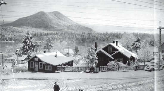Two dark-coloured houses beside each other while surrounded by snow. Behind them is a body of water, followed by a forest and a mountain. 
