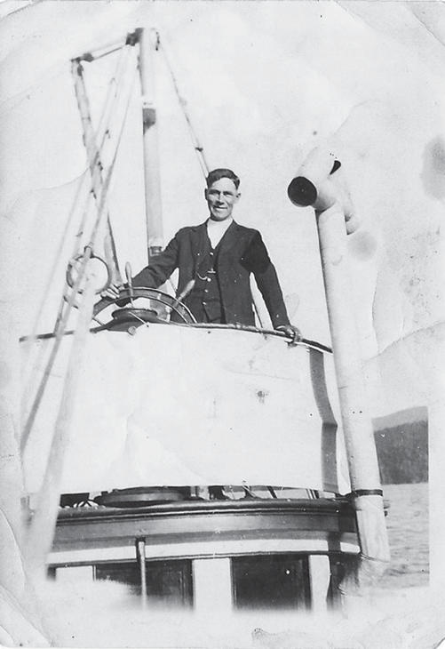 A person in a three-piece suit stands in the cockpit of a boat, their hand on the wheel while smiling at the camera.
