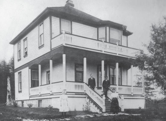 Four people stand in front of a two-storey, light-coloured house. 