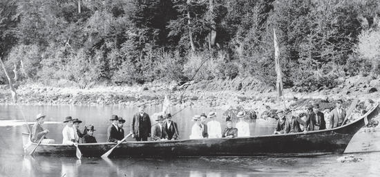 A docked canoe carrying about 19 people in hats and formal attire. Behind the canoe is a forest. 