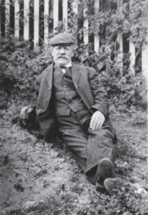 A man in a three-piece suit sits on a dirt-covered ground with his legs crossed while looking into the camera. The ground is at a decline. 