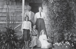 A man and woman standing in front of a building. In front of them are three younger girls, two sitting and one standing. They are all posed for the camera. 