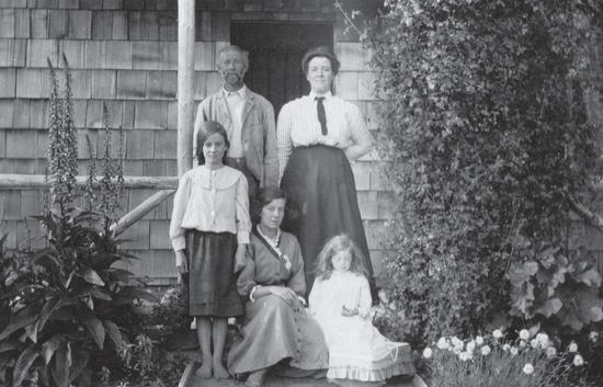 A man and woman standing in front of a building. In front of them are three younger girls, two sitting and one standing. They are all posed for the camera. 