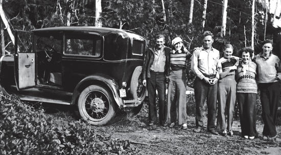 Six people pose together for the camera while standing next to an old dark-coloured car. Behind them is a forest. 