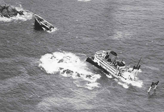 A large boat sinking into a body of water by a collection of rocks.