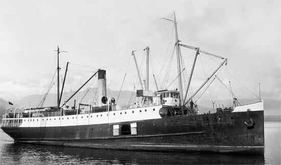 A very large steam ship in the water. The bottom of the ship is darker and the top is white. It has a tall stack in the center, as well as masts at either end. There are several cranes and pulleys on the ship. The name 'Princess Maquinna' is painted on the side near the front.