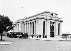 There is a railway station with vehicles parked on the left side of the building. There is a tree on the far right. Four people are standing in front of the building, and one is seated.