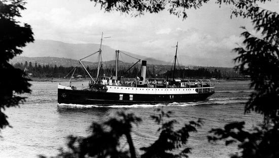 Photograph of the ship leaving Vancouver Harbour, framed by trees in the foreground, with the Northshore mountains in the background.