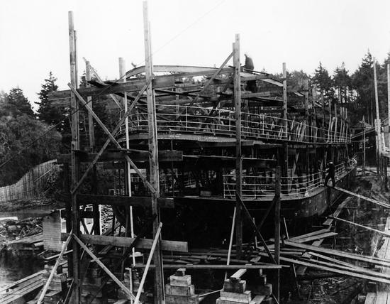 Photo depicts the ship  under construction at a dry dock, with wooden planks, piles, and a worker high up on the structure, backed by a forest.