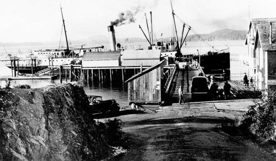 The photograph depicts one vehicle parked on the dock, one parked close to the pier's entrance, a few people near large buildings close to the port, and two structures on the pier closer to the Maquinna.