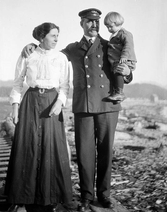 Captain Gillam, in uniform, holds a small child with blonde ear-length hair in one arm while his other is around a woman wearing a white blouse and a long dark skirt.