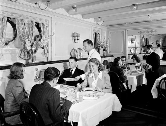 Promotional photo of the ships dining room: the scene shows formal table settings, flowers, white linens, formally dressed passengers smiling, and one white-jacketed waiter serving a dish to a man wearing a suit.