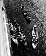 The photo depicts four rowboats from above, on a sunny day, loading passengers and supplies onto the Maquinna ship. Two of the four boats are rafted up, with men holding on, while the other two boats are out in the open water.