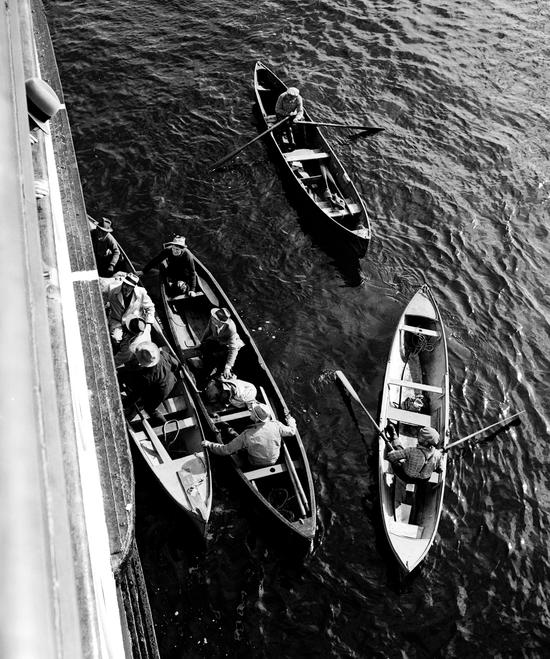 The photo depicts four rowboats from above, on a sunny day, loading passengers and supplies onto the Maquinna ship. Two of the four boats are rafted up, with men holding on, while the other two boats are out in the open water.