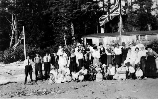 Group photo of British settlers on a beach with some seated with their children in the front. Two British flags, a forest and one building in the background.