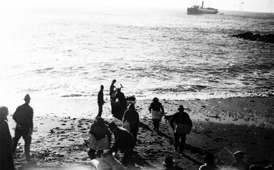 Photo of approximately thirteen people on shore landing supplies, a canoe near the shoreline with Princess Maquinna in the background.  