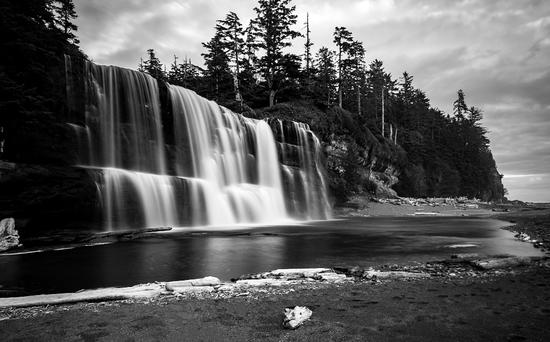 Photograph shows water cascading gently over rocks on the left, with trees in the background and a sandy beach on the right. 