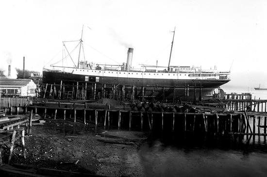 The photo displays the ship's side view while it's at dry dock. Logs and a small sandy beach are visible in the foreground. 