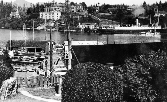 Photo of the ship in the distance, smokestack steaming while docked at the telegraph station across the inlet in Bamfield. The image also captures various buildings, docks, and houses.