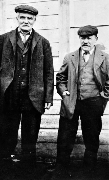 Two men dressed in suits and caps both with moustaches. 