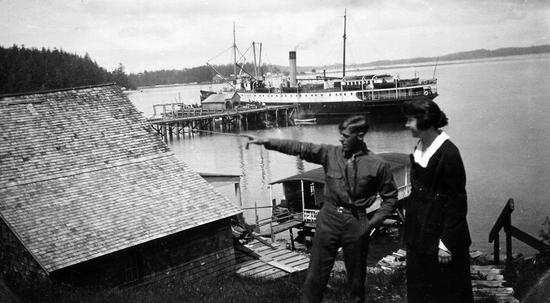 Harold Monks and Margery McCannel stand near a dock in Tofino, with the Princess Maquinna docked behind them. Harold points to his right, Margery watches.