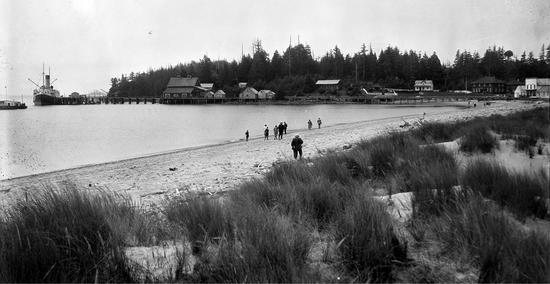 Photo of people walking on the beach with a ship visible in the distance and beach grass in the foreground.
