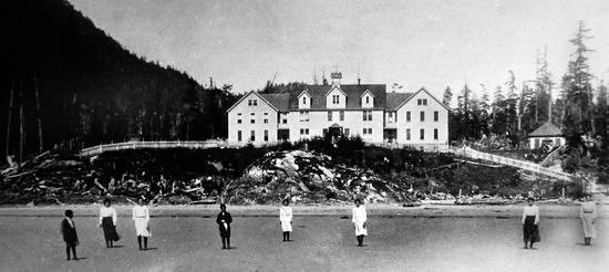 Eight boys and girls stand far apart on a windy day, facing the camera on a sandy beach with the Christie Residential School in the background.