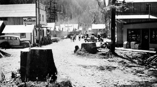 This photo shows snow is on the ground in a small town with buildings and cars from the 1940s. People are walking in the distance, and a forest is in the background.