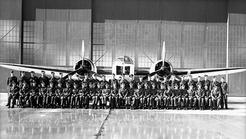 Formal photographic portrait of air crew, sitting and standing in front of a Bolingbroke bomber aircraft, inside a hangar.