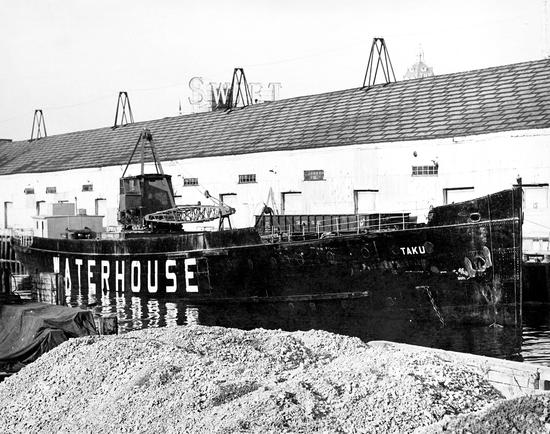 Photograph of the Princess Maquinna next to a building in the water as the Taku, an ore barge.
