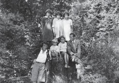 A family photo of three girls, two boys, and their mother and father, all standing before a backdrop of lush forest