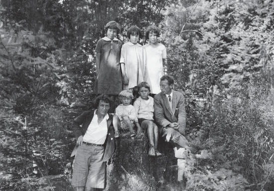 A family photo of three girls, two boys, and their mother and father, all standing before a backdrop of lush forest
